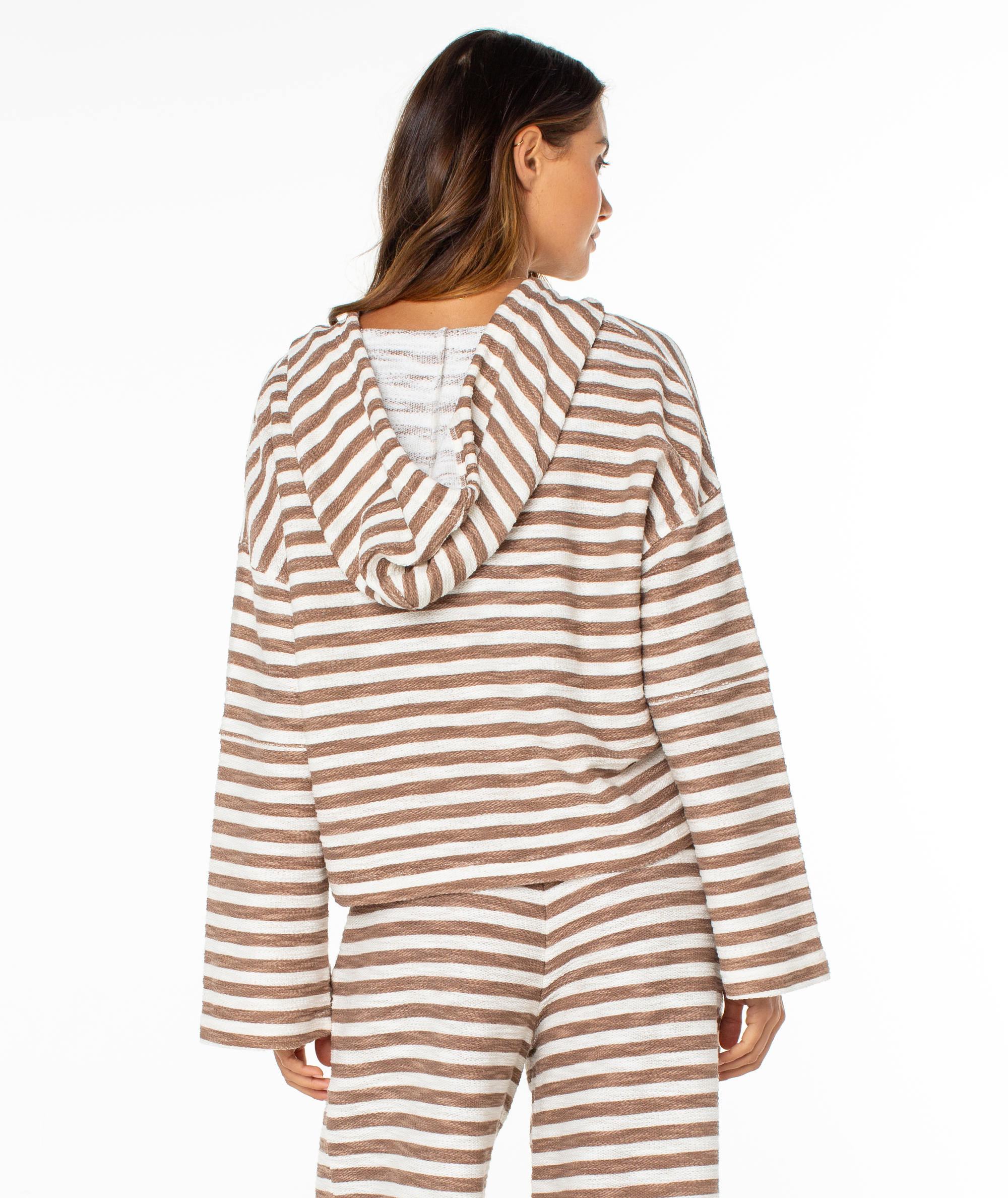 A woman is seen from behind wearing the Roxy Rich Coast Stripe Hoodie in brown and white. Made from soft terry fabric, it exudes cozy, beachy vibes as she stands against a plain white background.