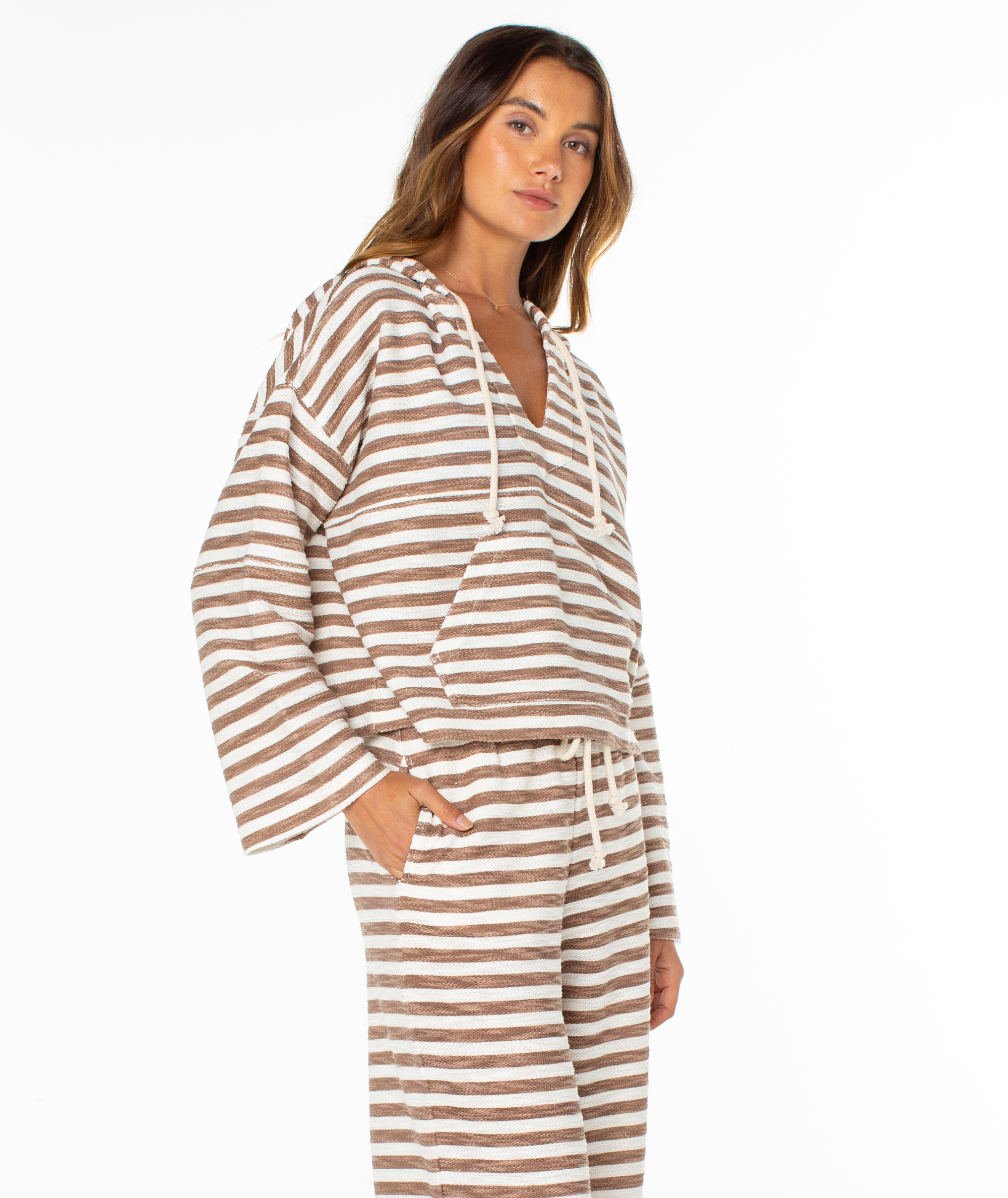 A woman with long brown hair and a neutral expression wears the Roxy Rich Coast Stripe Hoodie—a loose, brown and white striped terry top with drawstrings—paired with matching pants for easy, beach-inspired style.