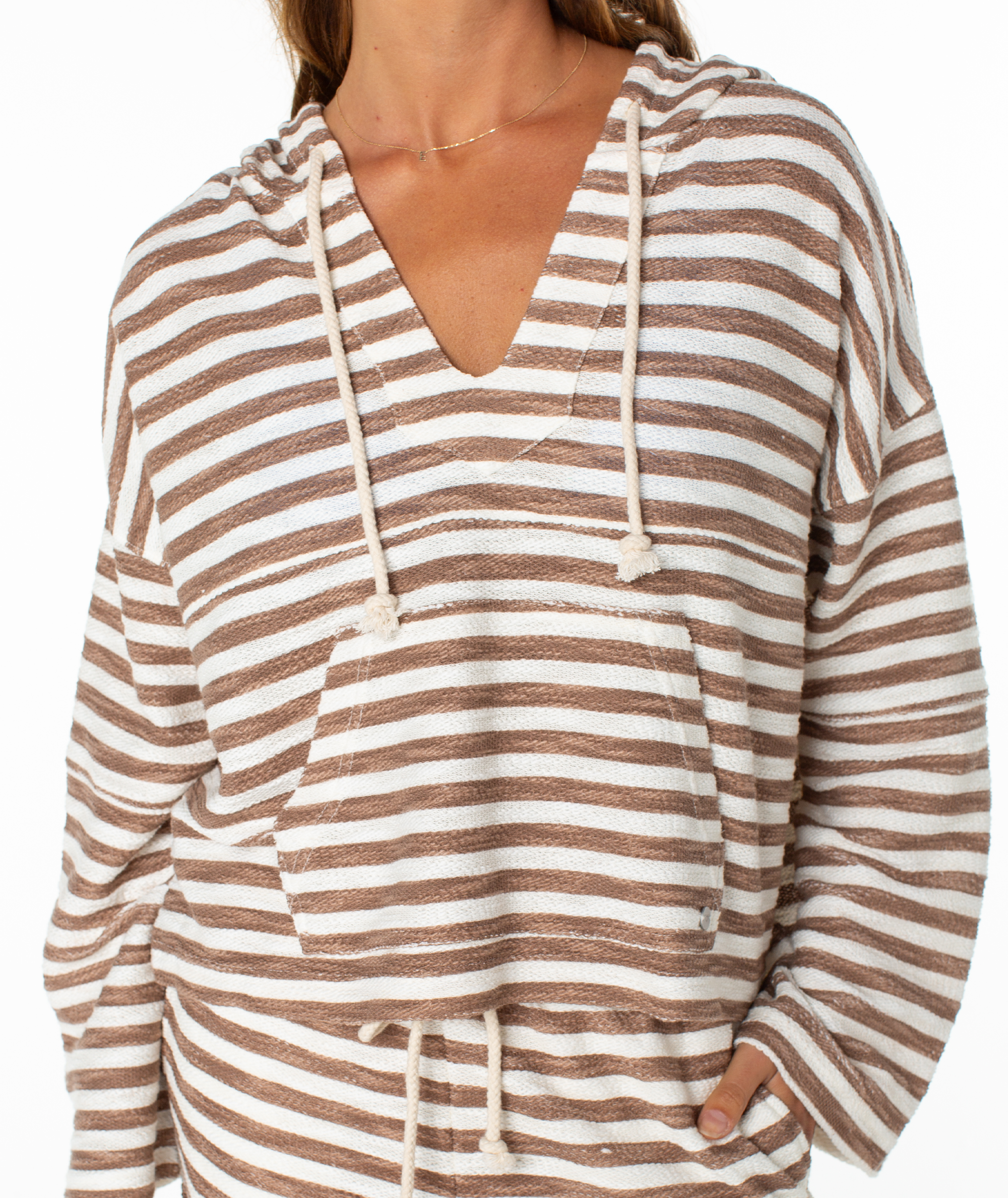 A person stands with one hand in the pocket of a Roxy Rich Coast Stripe Hoodie, featuring brown and white stripes, front pocket, and drawstrings. Cozy terry fabric adds a beachy vibe. Face is partially out of frame.