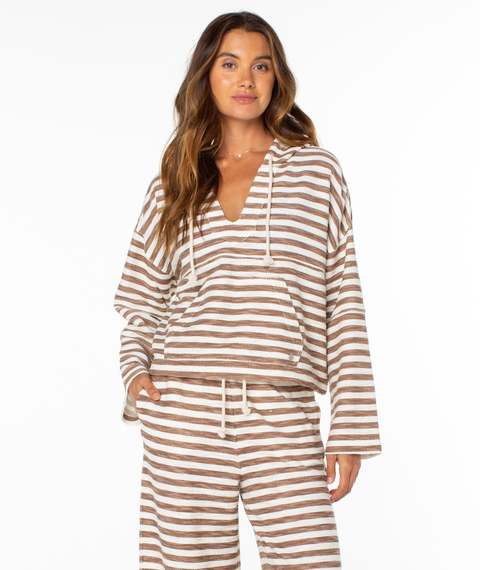 A woman with long brown hair wears the Roxy Rich Coast Stripe Hoodie—a tan and white striped top with a hood and front pouch—paired with drawstring pants. She stands against a plain white background, looking ahead and smiling slightly.