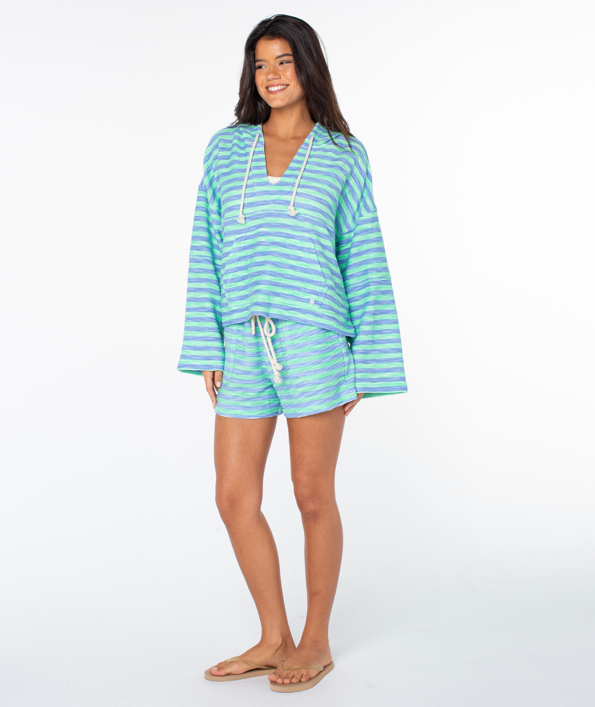 A woman smiles, wearing a loose, long-sleeved Roxy Rich Coast Stripe Hoodie in green and white with matching terry shorts and tan flip-flops. She stands against a plain white background.