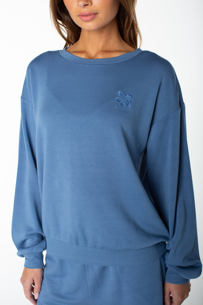 A woman wears the Roxy Surfing By Moonlight Top, a loose blue pullover with a subtle embroidered logo and matching bottoms, crafted from soft modal stretch fabric, standing against a plain white background.