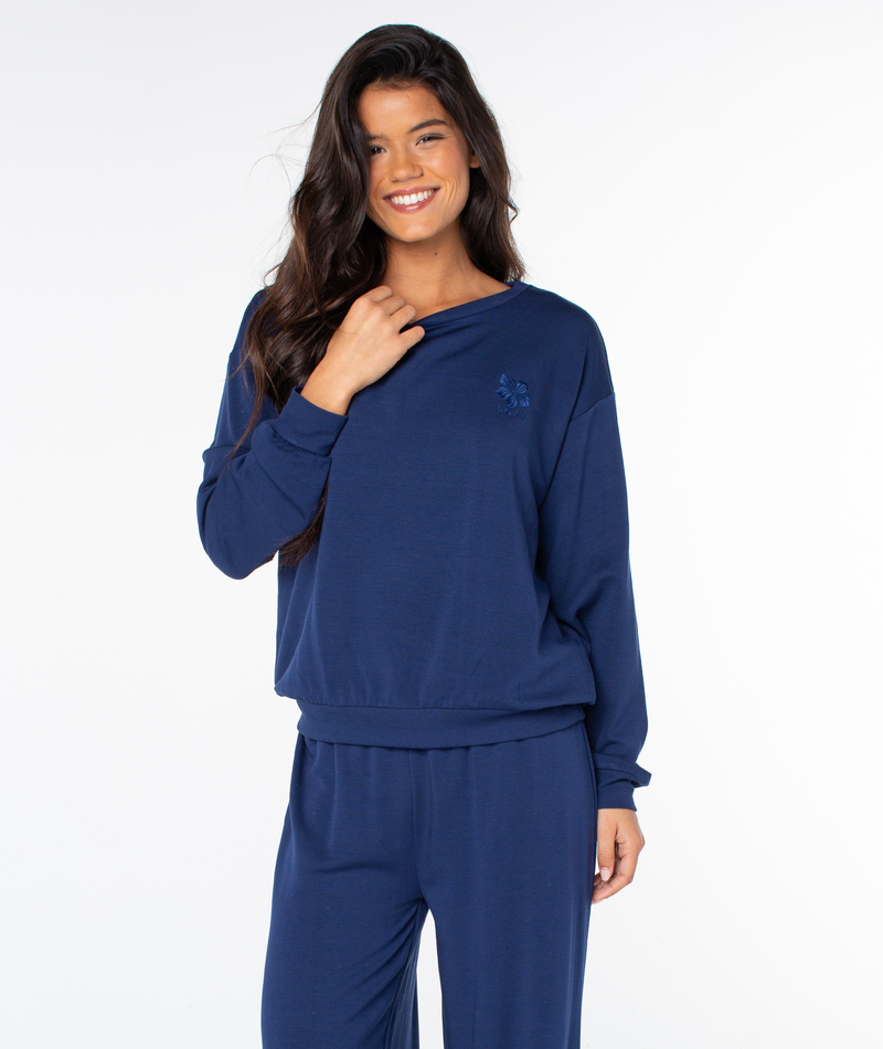 A woman smiles wearing a cozy, long-sleeved navy blue Roxy Surfing By Moonlight top with a small flower emblem on the chest.