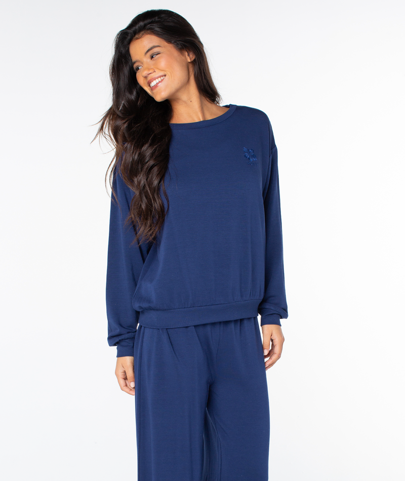 A smiling woman wearing a navy blue Roxy Surfing By Moonlight Top, a long-sleeved crewneck sweatshirt with a small embroidered logo on the chest.