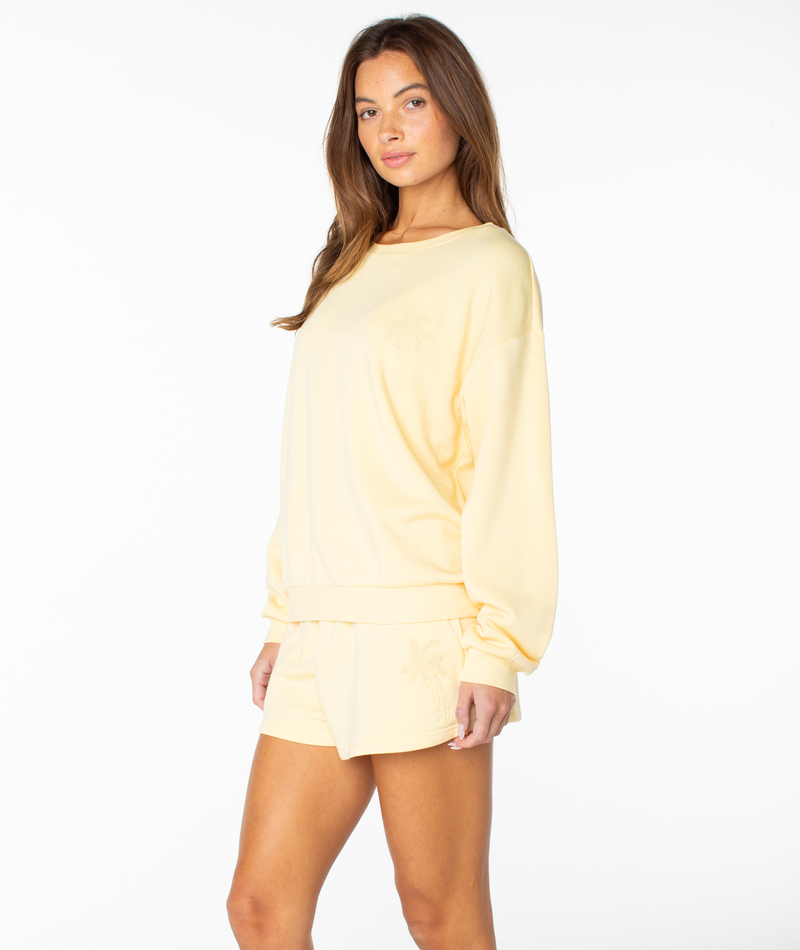 A woman stands against a white background, slightly turned and looking at the camera, wearing the Roxy Surfing By Moonlight Top—a cozy light yellow pullover with matching palm tree shorts that add easy comfort to her style.