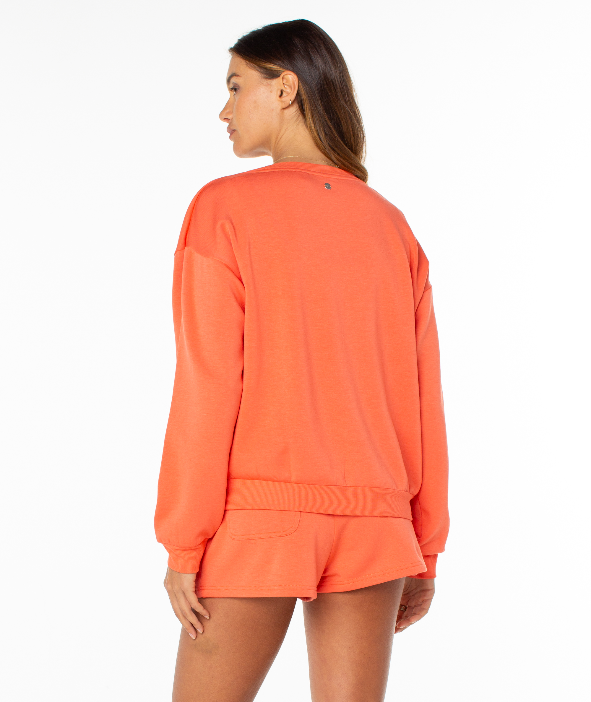 A woman stands with her back turned, wearing the Roxy Surfing By Moonlight Top—an orange, cozy long-sleeve pullover made from modal stretch fabric—against a plain white background.