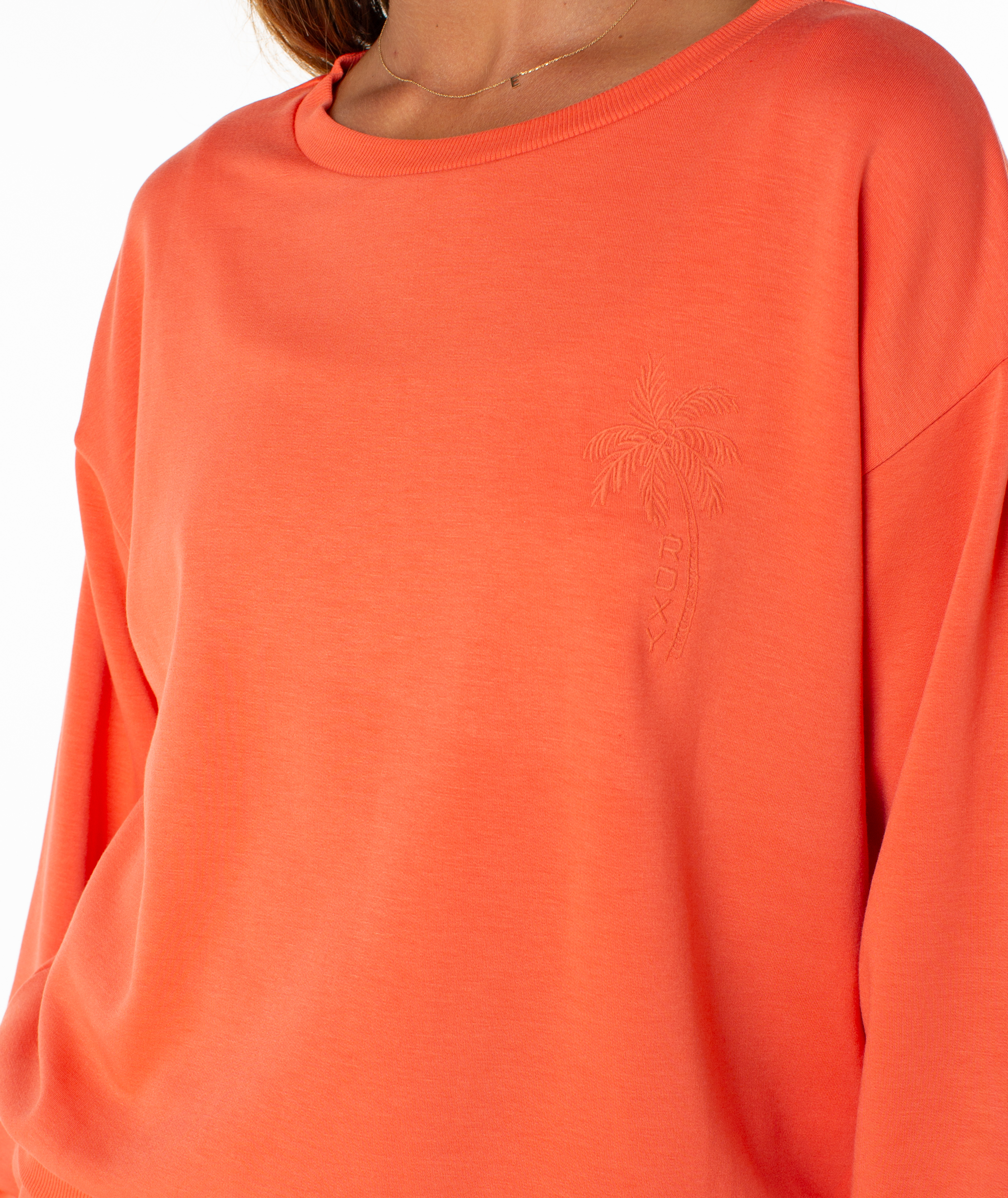 A close-up of a woman wearing the Surfing By Moonlight Top by Roxy, an orange crewneck fleece featuring an embroidered palm tree and ROXY logo on the left chest.