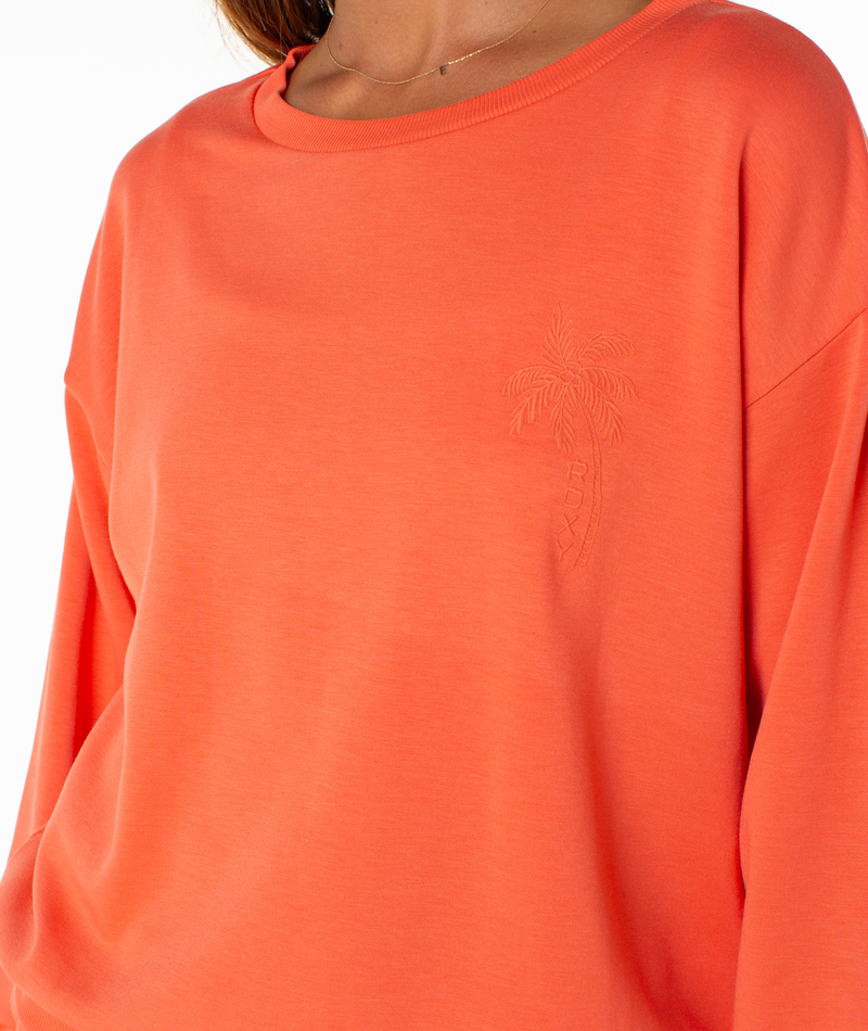 A close-up of a woman wearing the Surfing By Moonlight Top by Roxy, an orange crewneck fleece featuring an embroidered palm tree and ROXY logo on the left chest.