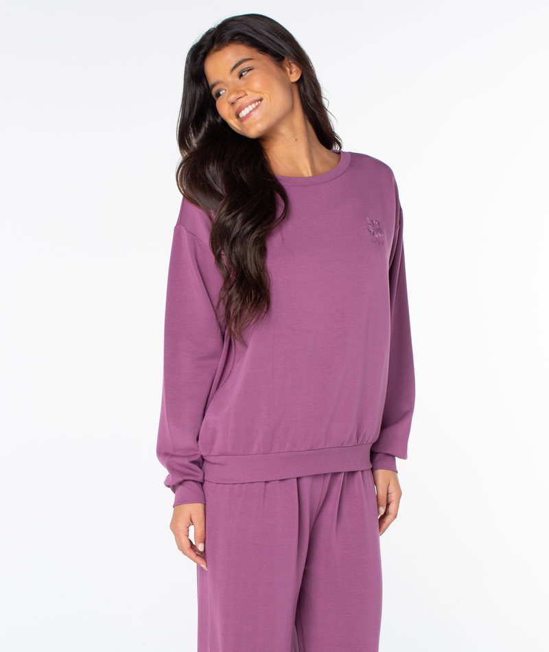 A smiling woman wearing a cozy, purple long-sleeve crewneck Surfing By Moonlight Top by Roxy.