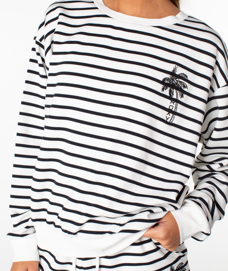 Model is wearing the Roxy Surfing By Moonlight Stripe Top—a cozy white pullover sweatshirt with black horizontal stripes and a small embroidered palm tree on the left chest—standing against a white background, showcasing a relaxed look that perfectly captures classic beachwear style.