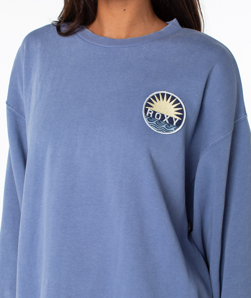 Person wears a Hazy Daze Patch Top by Roxy in blue—a cozy cotton blend with a round ROXY patch depicting a sunrise over waves. Only the upper body is shown against a plain white background.