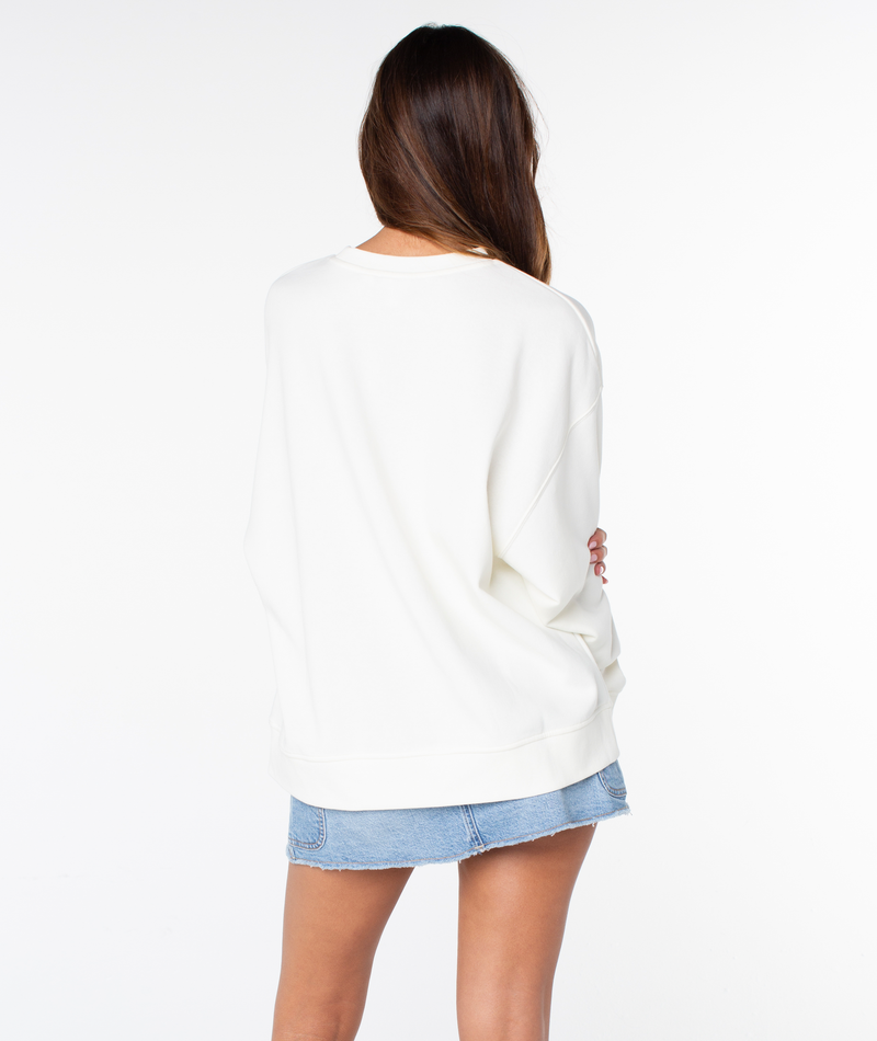 A woman with long brown hair faces away, wearing the Roxy Morning Stroll Top—a cozy cotton crewneck white sweater—and a short denim skirt against a plain white background.