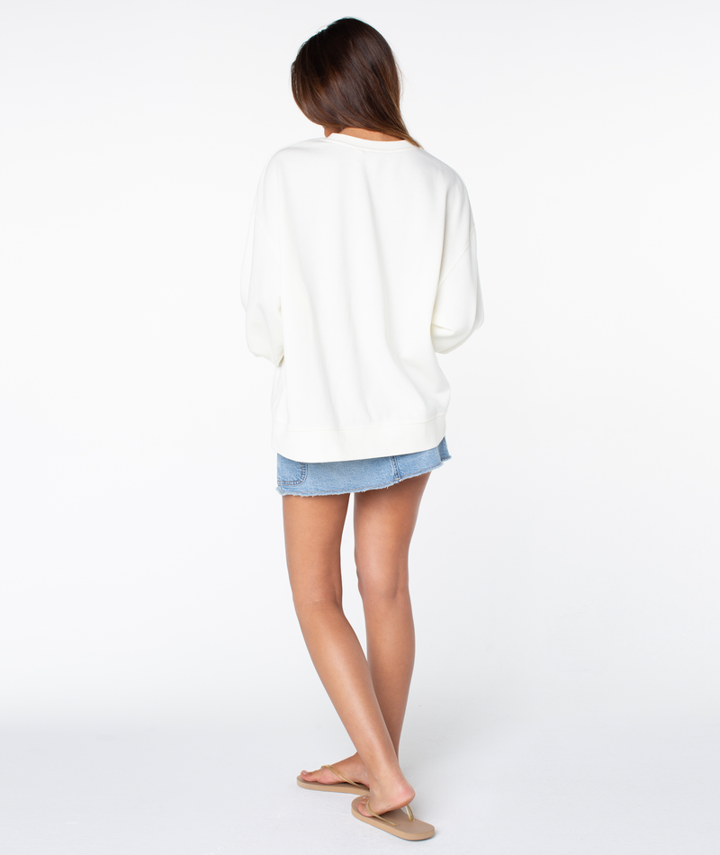 A person faces away, wearing the Roxy Morning Stroll Top— a cozy cotton crewneck white long-sleeve—paired with a denim skirt and beige sandals for an effortless, everyday style against a white background.