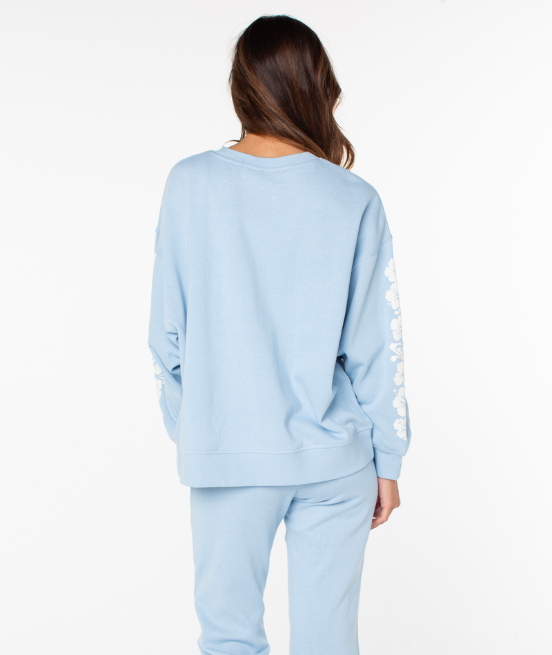 A person with long brown hair wears the Roxy Morning Stroll Top, a cozy light blue cotton crewneck shown from the back, featuring matching pants and white floral designs on the sleeves.