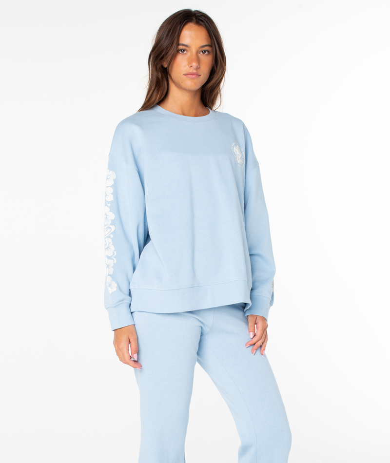 A person stands against a white background wearing the Roxy Morning Stroll Top in Cerulean Vintage Hibiscus, featuring cozy cotton fabric and white floral designs on the sleeves, paired with matching pants.