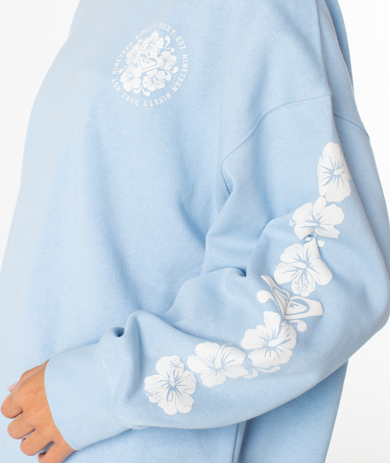 A person wearing the Morning Stroll Top by Roxy—a cozy cotton crewneck in light blue with white floral sleeve designs and a small circular floral graphic on the chest.