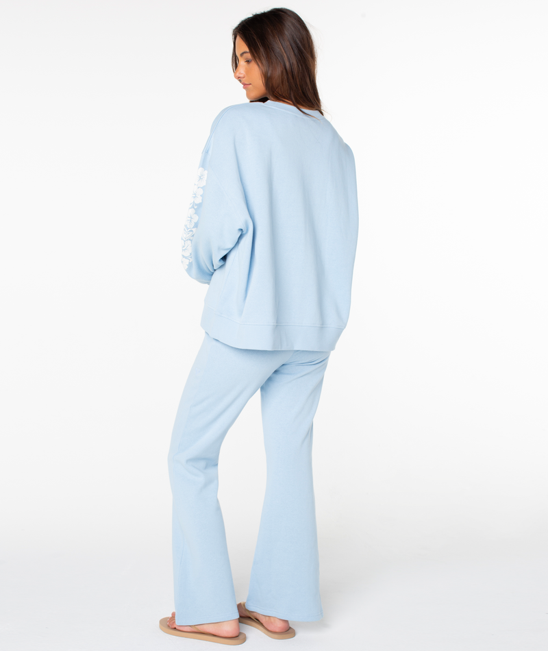 A woman stands on a plain white background, glancing over her shoulder while wearing the Roxy Morning Stroll Top with flared pants and flip-flops for an effortlessly cozy look.