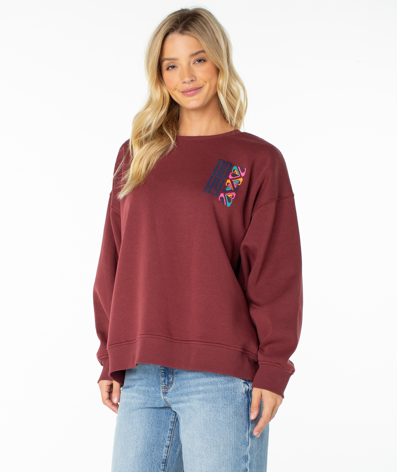 A woman with long blonde hair is standing and smiling against a plain white background, wearing the Roxy Morning Stroll Top in Port Oceanview—a beachy-cool maroon graphic crewneck sweatshirt featuring a colorful embroidered chest design—paired with light blue jeans.