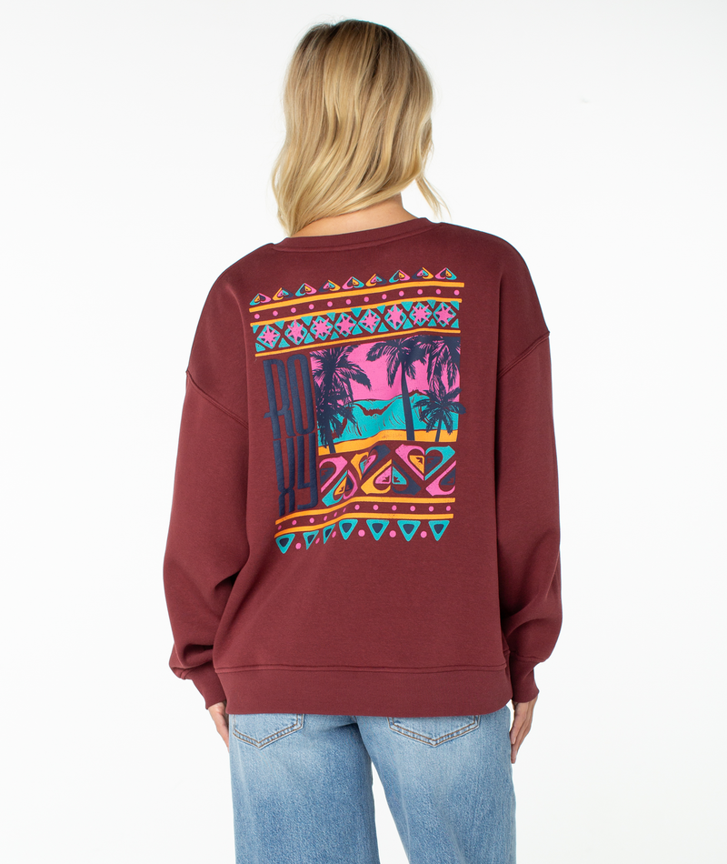 Wearing the Roxy Morning Stroll Top, a maroon graphic crewneck sweatshirt with a vibrant tropical beach and palm tree design on the back, a blonde-haired person pairs it with light blue jeans for an effortlessly laid-back, beachy-cool vibe.