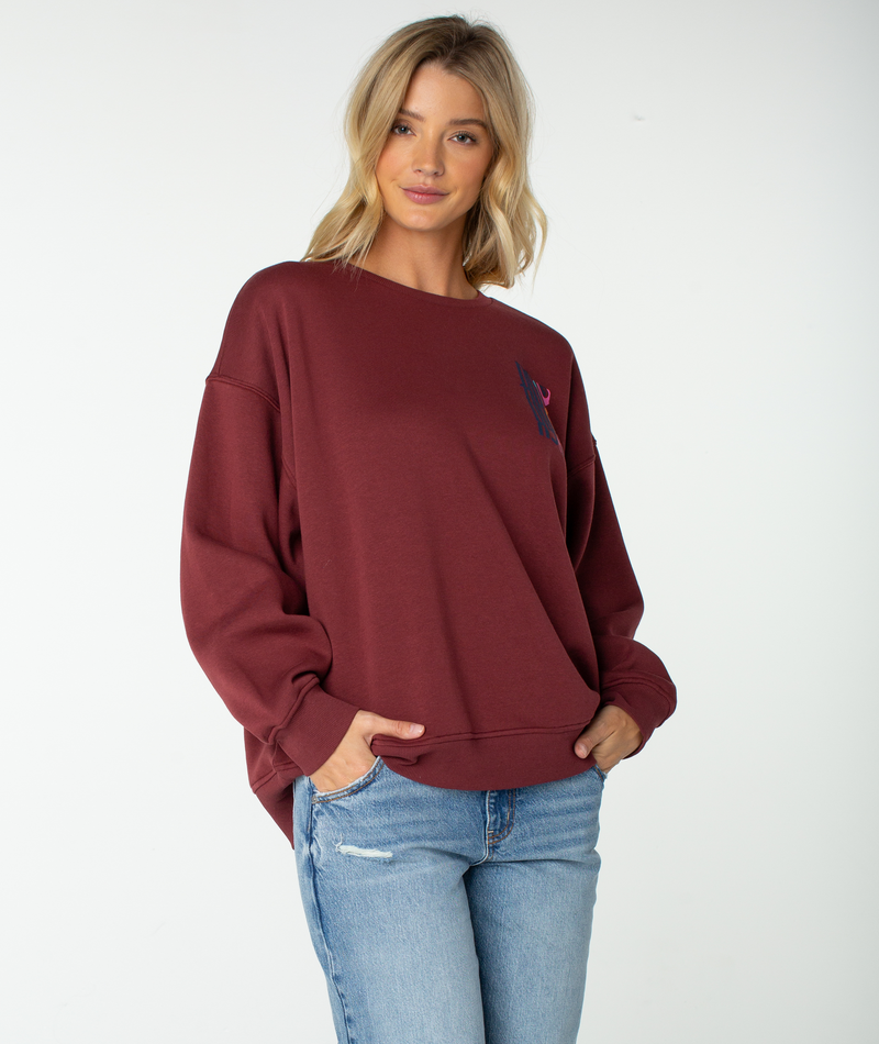 A woman with blonde hair wears the Roxy Morning Stroll Top, a cozy maroon fleece sweatshirt, paired with light blue jeans. She stands against a plain white background with her hands in her pockets, smiling gently at the camera.