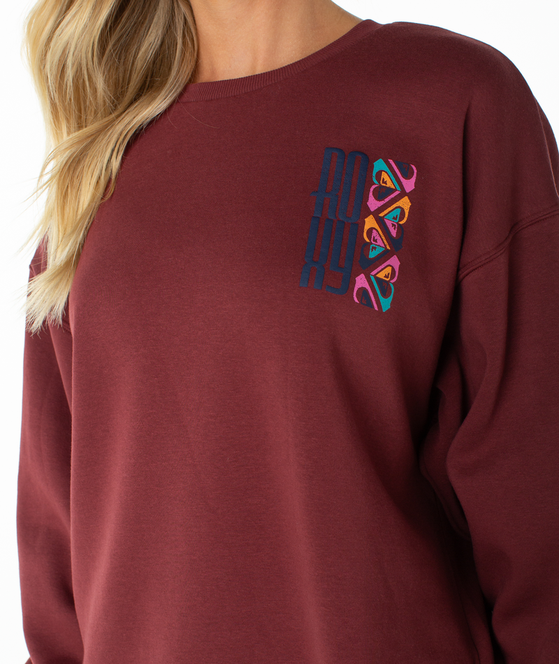 A person is wearing the Roxy Morning Stroll Top, a maroon graphic crewneck sweatshirt featuring colorful embroidered details and “ROXY” across the chest. Shown from the shoulders down with long blonde hair, the look captures a laid-back, beachy-cool vibe.