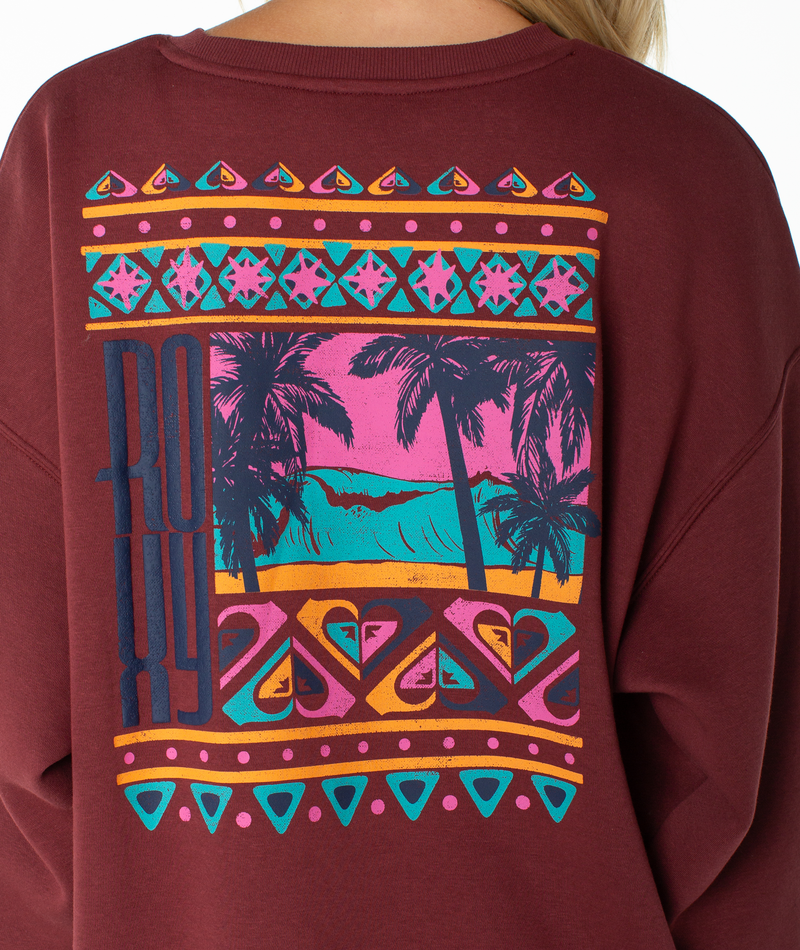 Wearing the Roxy Morning Stroll Top, a maroon graphic crewneck sweatshirt featuring a vibrant tropical beach scene with palm trees and geometric patterns on the back, plus bold ROXY lettering on the left side for an effortlessly beachy-cool vibe.