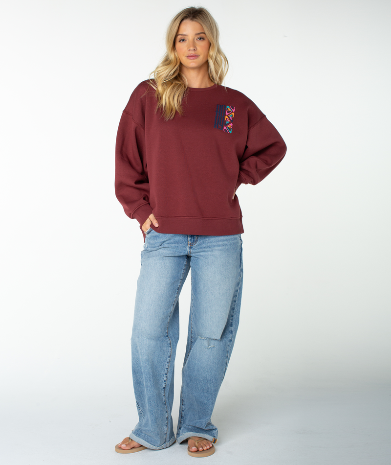 A person stands against a white background wearing the Roxy Morning Stroll Top—a maroon graphic crewneck with colorful details—paired with light blue jeans and sandals, one hand on their hip and the other relaxed.