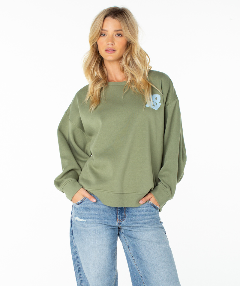 A woman stands with her hands in her pockets facing forward against a plain white background, wearing the Roxy Morning Stroll Top in Oil Green Wave Riders Club and light blue jeans.