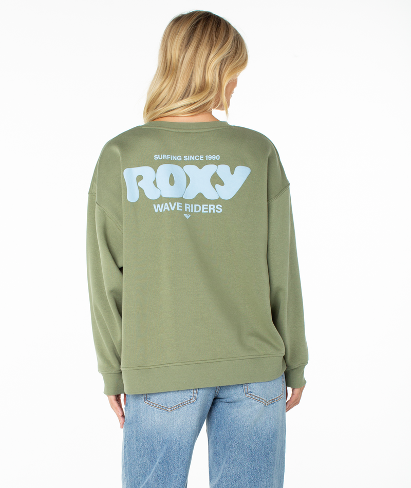 A woman with blonde hair is seen from behind wearing blue jeans and the Roxy Morning Stroll Top, a green graphic crewneck sweatshirt. The back of the sweatshirt showcases large, light blue text that reads “SURFING SINCE 1990 ROXY WAVE RIDERS,” adding a casual, beachy-cool vibe.