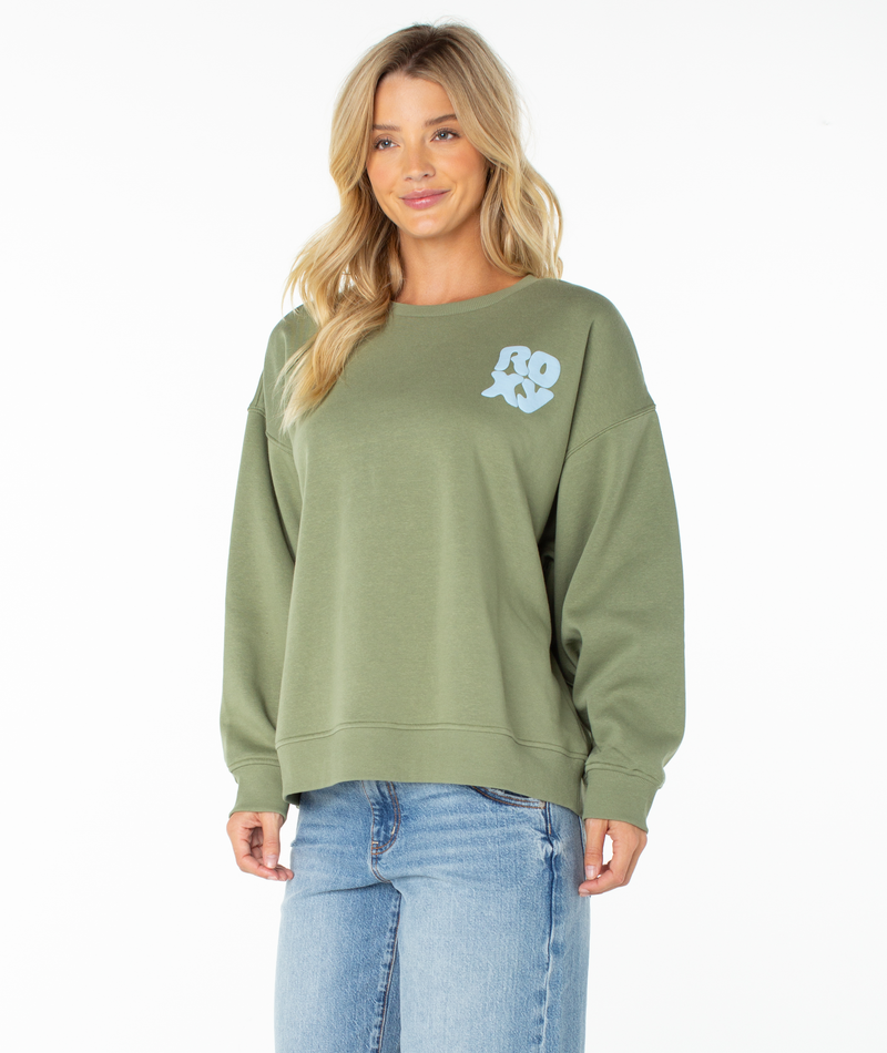 A woman wearing the Roxy Morning Stroll Top, a sage green graphic crewneck sweatshirt with “ROXY” printed in white on the chest, pairs it with light blue jeans and poses against a plain white background for a laid-back, beachy vibe.