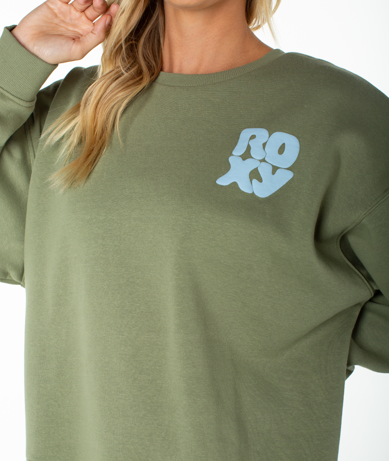 A person wearing the Roxy Morning Stroll Top, a cozy olive green fleece sweatshirt featuring ‘ROXY’ in light blue lettering on the left chest. With their face partially out of frame and a hand tucked into their hair, they embody an effortless beachy-cool vibe.