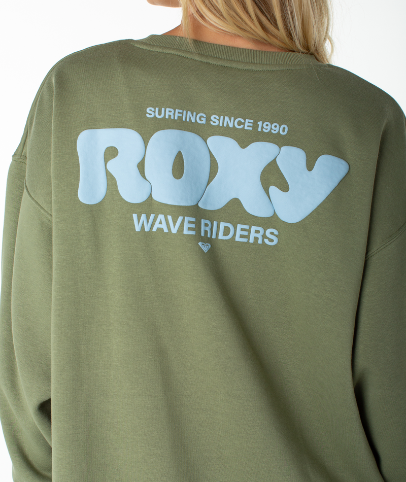 A person wearing the Roxy Morning Stroll Top, an olive green graphic crewneck sweatshirt with blue text on the back that reads “ROXY, SURFING SINCE 1990, WAVE RIDERS.”.