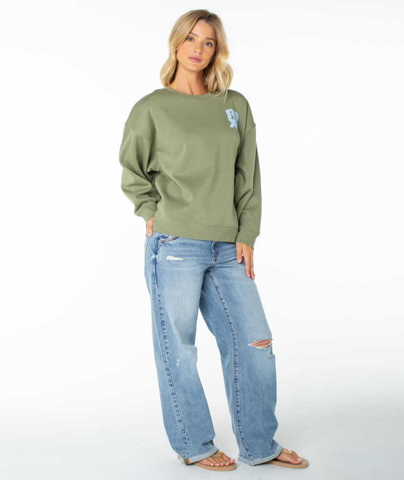 A woman with blond hair poses in a relaxed, beachy-cool style against a white background, wearing the Roxy Morning Stroll Top—an olive green graphic crewneck sweatshirt with a blue design—paired with ripped light blue jeans and sandals.