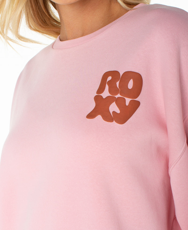 A person wearing the Roxy Morning Stroll Top, a light pink beachy crewneck sweatshirt featuring the word “ROXY” in bold, wavy brown letters across the chest. Only the lower face, neck, and upper torso are shown.