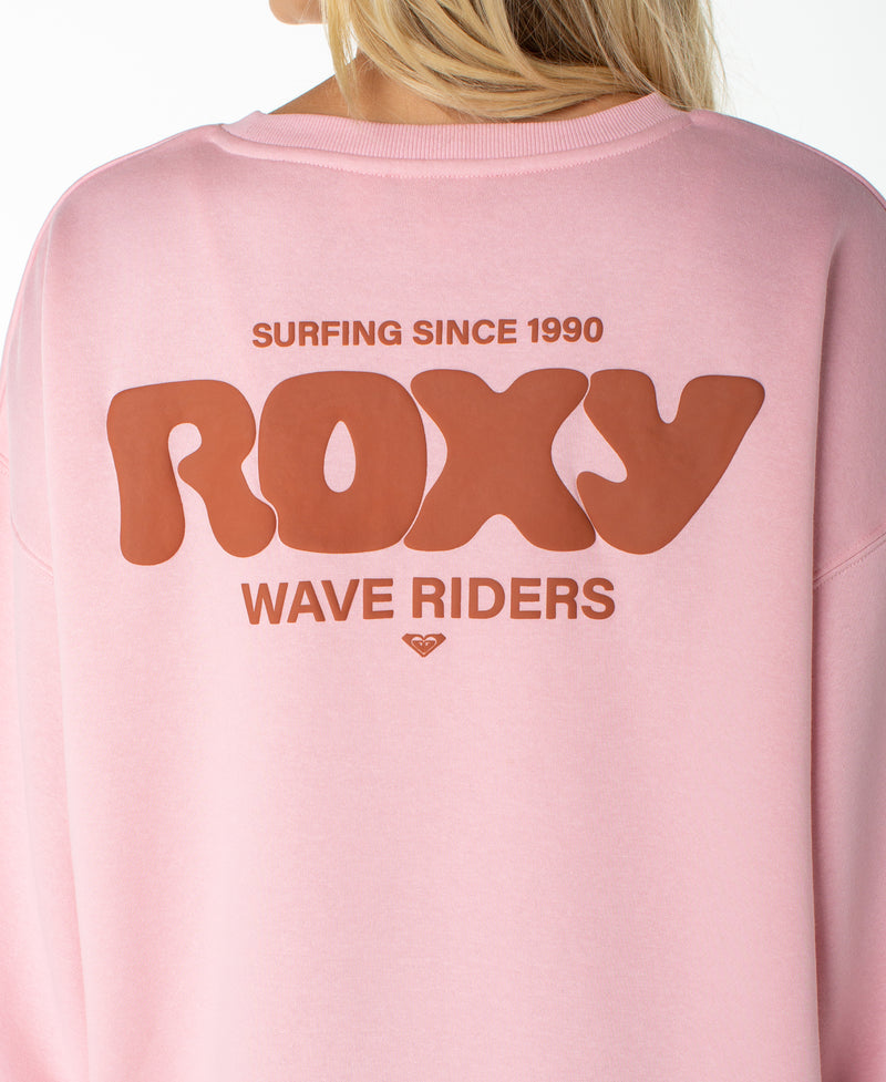 A person wearing the Roxy Morning Stroll Top, a pink graphic crewneck sweatshirt featuring bold “Surfing Since 1990, ROXY, Wave Riders” text on the back.