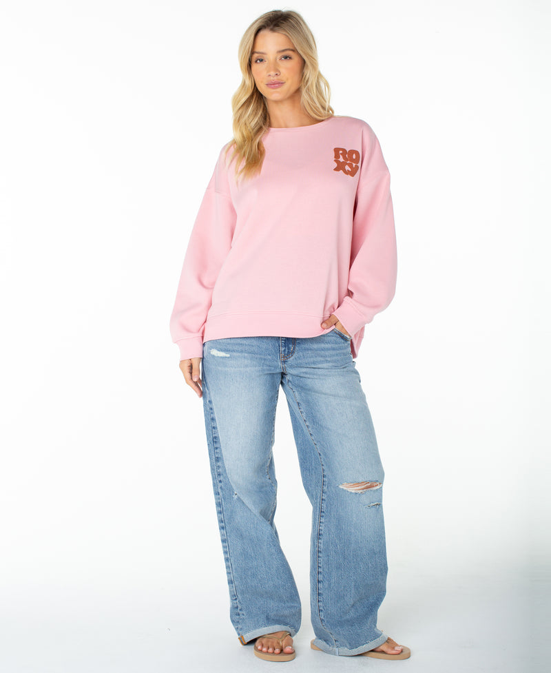 A woman stands facing forward against a plain white background, wearing the Roxy Morning Stroll Top—a cozy light pink fleece graphic crewneck sweatshirt with “ROXY” displayed on the chest—paired with light blue ripped jeans. She has long blonde hair and is barefoot.