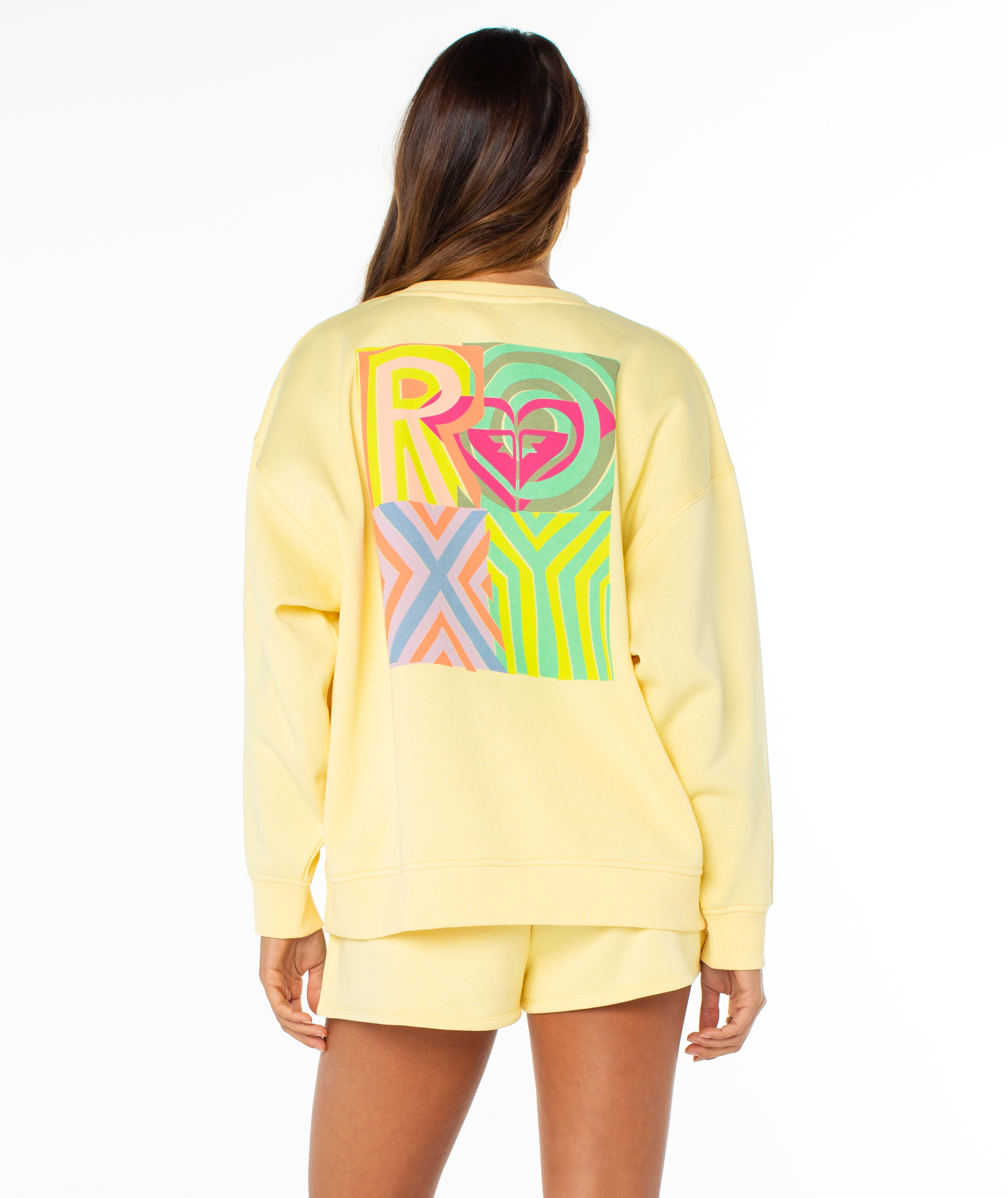 A woman with long brown hair wears the Morning Stroll Top by Roxy, a light yellow graphic crewneck sweatshirt with a colorful abstract print on the back. She pairs it with matching shorts and stands against a white background.