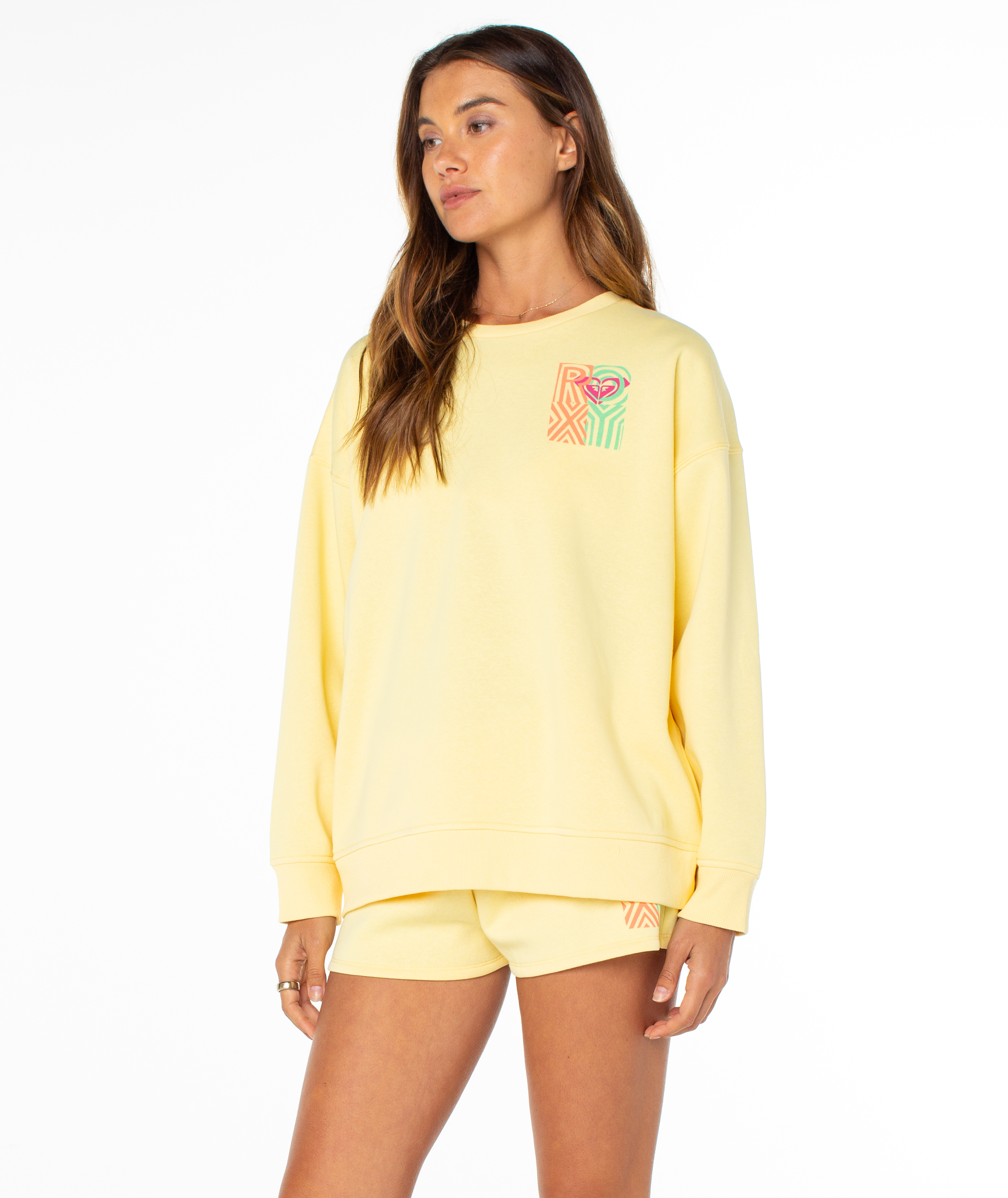 A woman wears the Roxy Morning Stroll Top, a loose-fitting pale yellow graphic crewneck sweatshirt with a colorful abstract design, standing against a plain white background—a perfect summer staple.