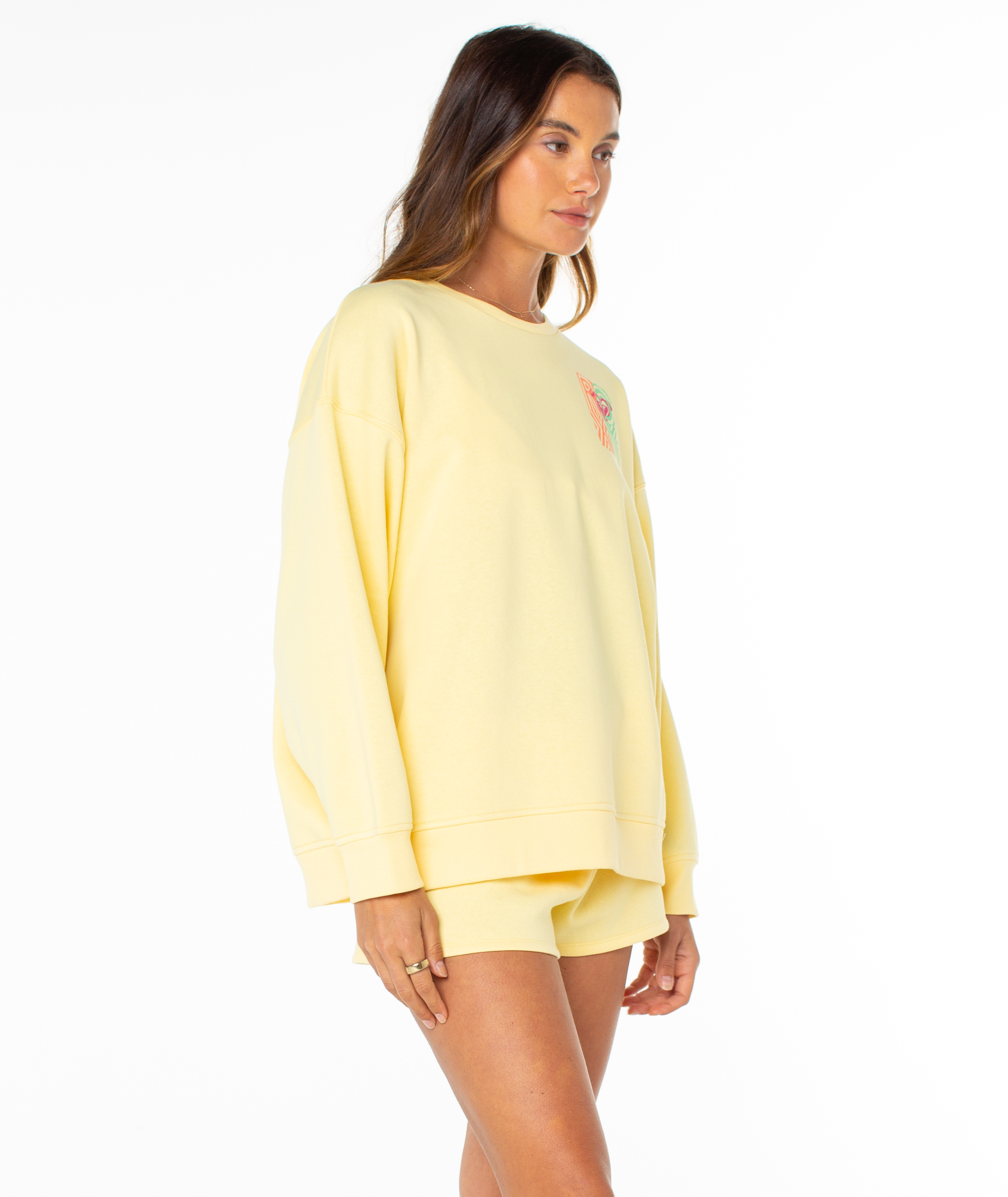 A woman stands sideways in the Roxy Morning Stroll Top, a loose-fitting pastel yellow graphic crewneck. The cozy fleece sweatshirt offers casual comfort for summer, shown here against a white background.