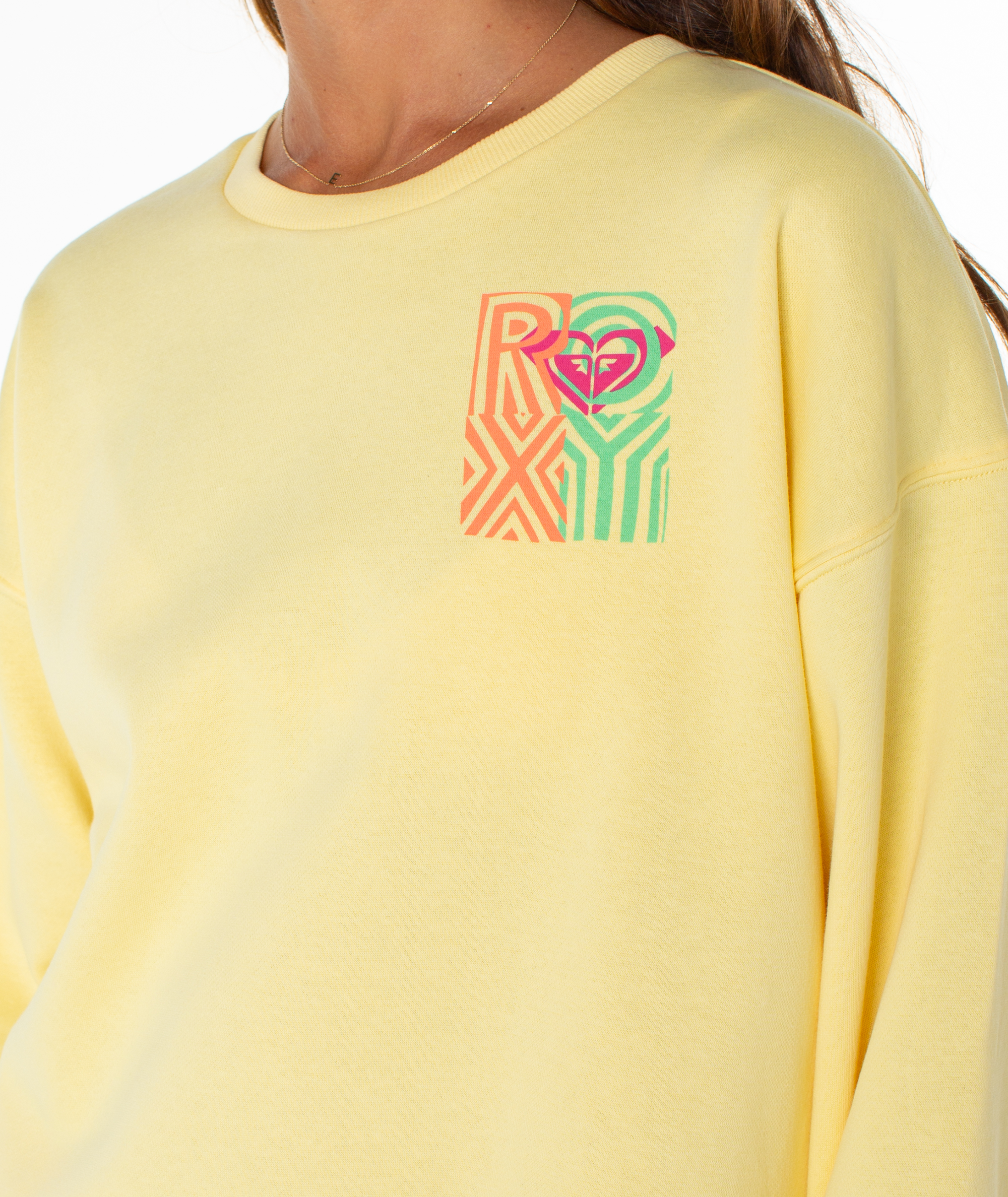 A person wearing the Roxy Morning Stroll Top, a light yellow crewneck sweatshirt featuring a colorful ROXY logo and heart graphic on the upper left chest. Only their lower face, neck, and upper body are visible.