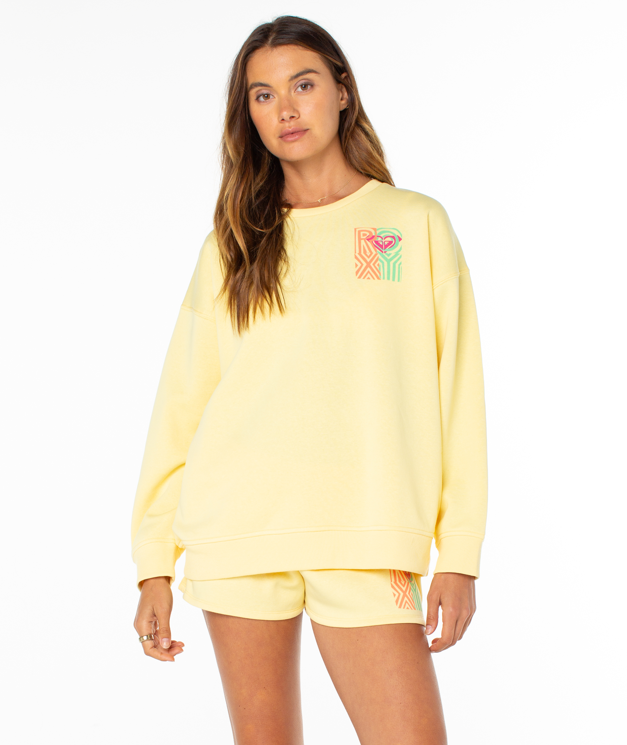 A woman with long hair poses against a white background in the Roxy Morning Stroll Top, a loose-fitting pale yellow crewneck sweatshirt featuring colorful geometric graphics—perfect as a cozy summer staple.