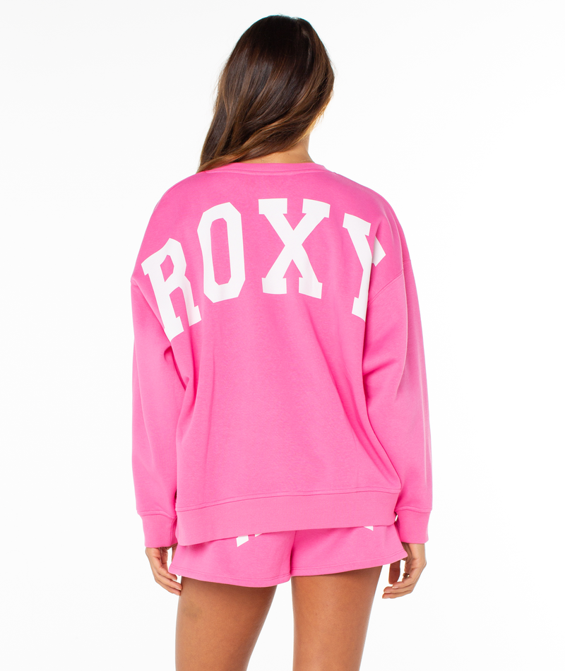 A person with long brown hair wears the Surfing By Moonlight Top by Roxy, a cozy bright pink fleece sweatshirt with large white “ROXY” lettering on the back. Matching shorts complete the look against a plain white background.