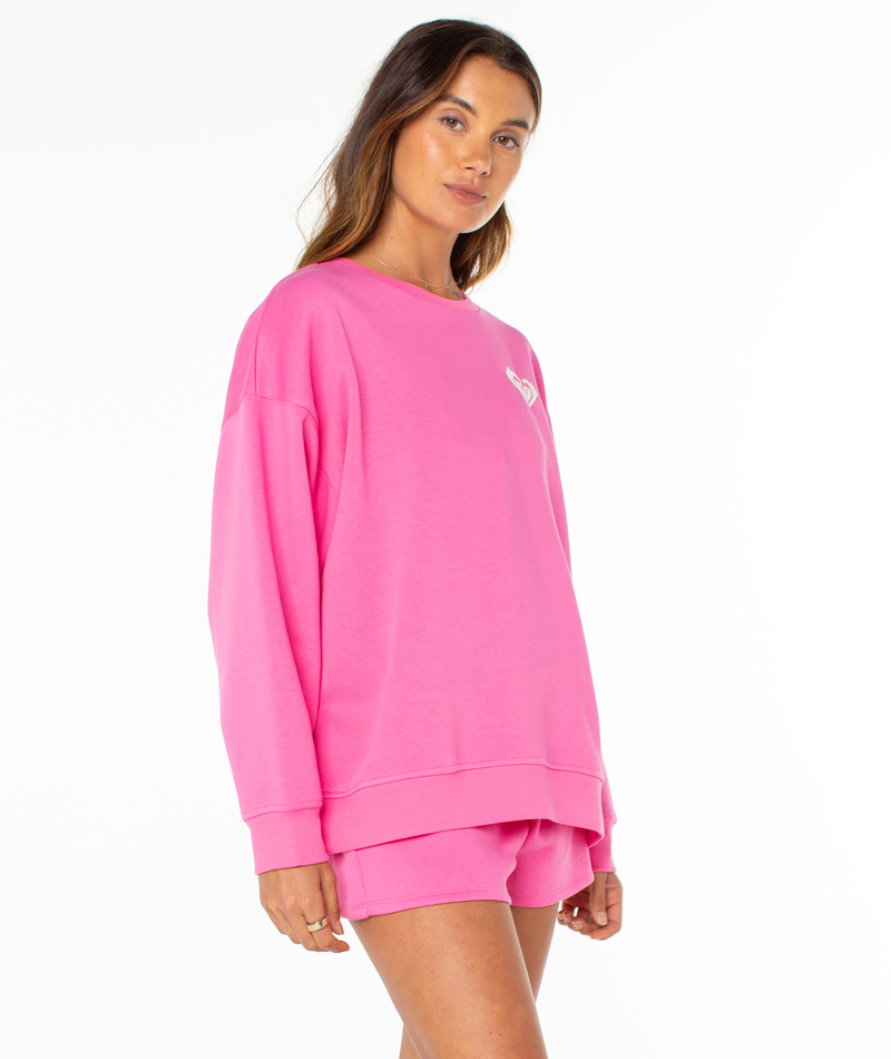 A woman stands against a white background wearing the Roxy Surfing By Moonlight Top, a bright pink graphic crewneck sweatshirt, paired with matching pink shorts and looking at the camera with a slight smile.