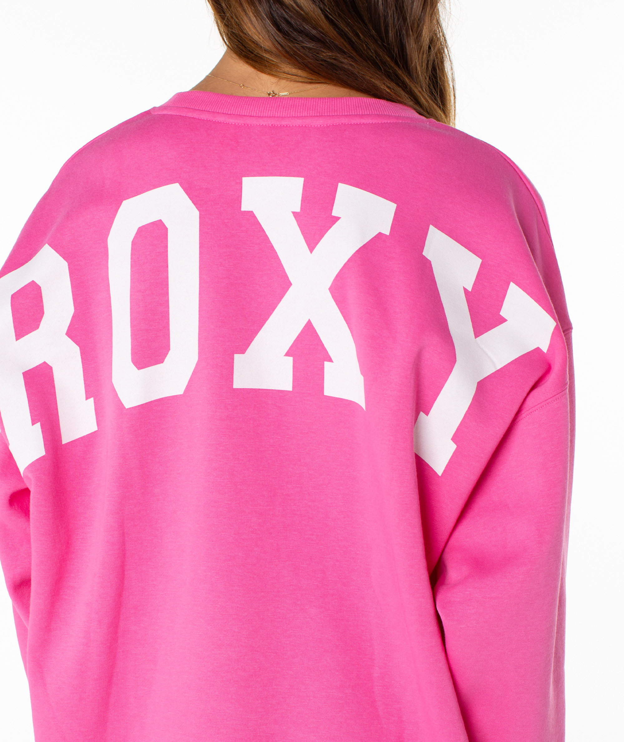 A person is seen from behind, wearing the Roxy Surfing By Moonlight Top—a bright pink graphic crewneck with bold white "ROXY" lettering on the back and only their upper back and brown hair partially visible.