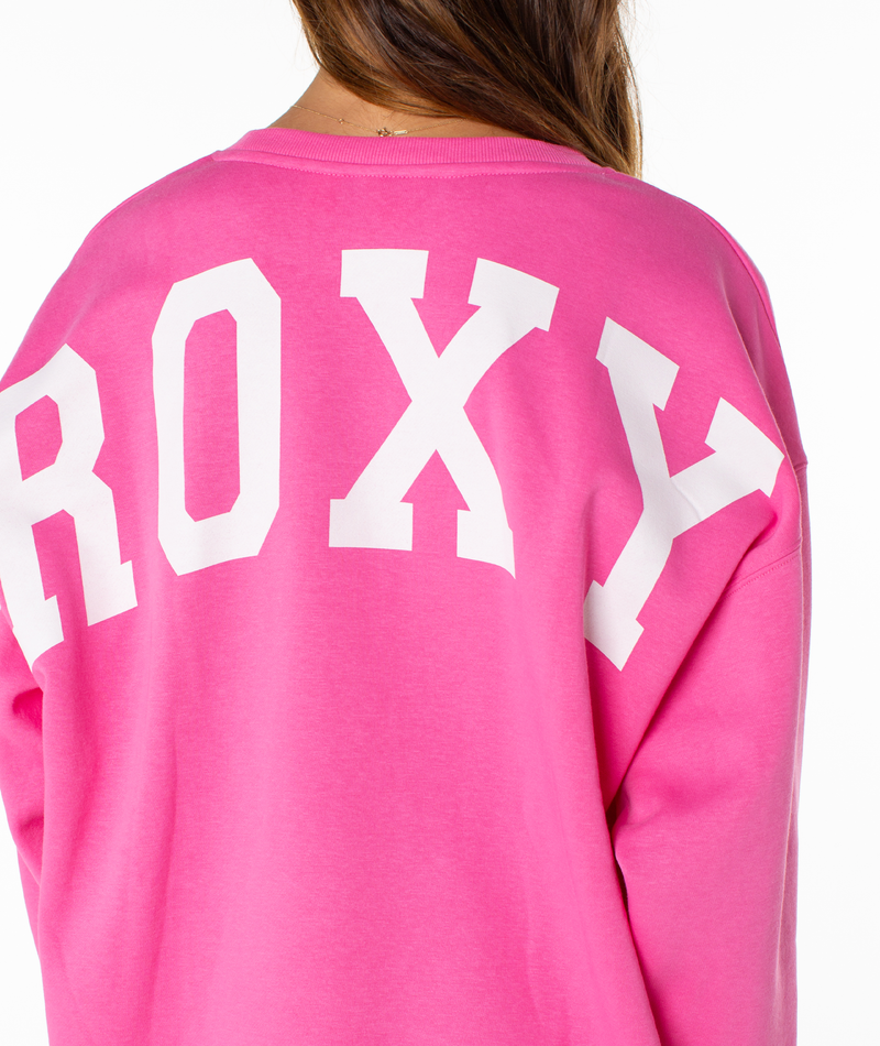 A person is seen from behind, wearing the Roxy Surfing By Moonlight Top—a bright pink graphic crewneck with bold white "ROXY" lettering on the back and only their upper back and brown hair partially visible.