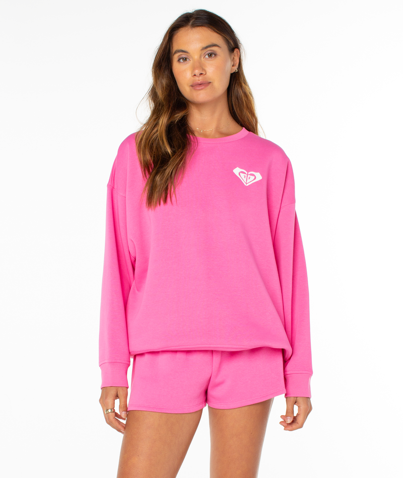 A woman with long brown hair and a relaxed expression wears the Roxy Surfing By Moonlight Top, a bright pink graphic crewneck sweatshirt with a small white logo on the left chest, paired with matching shorts against a plain white background.