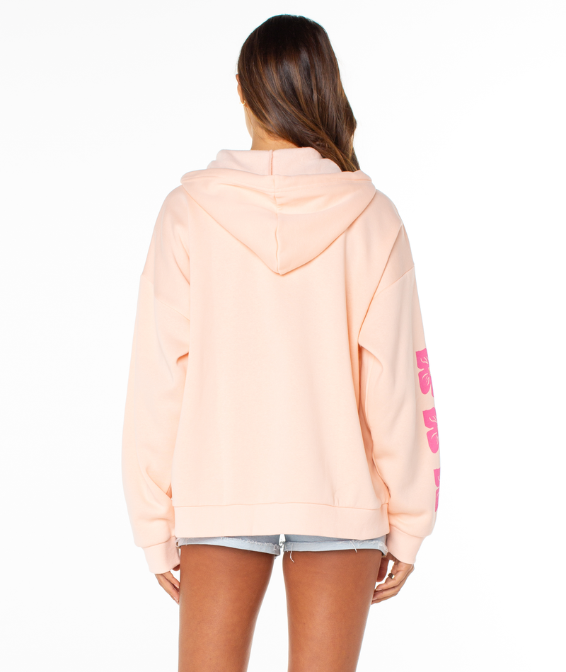 A woman with long brown hair wears the Roxy Evening Lineup Hoodie in light peach with pink sleeve lettering and light denim shorts, standing facing away from the camera against a white background.