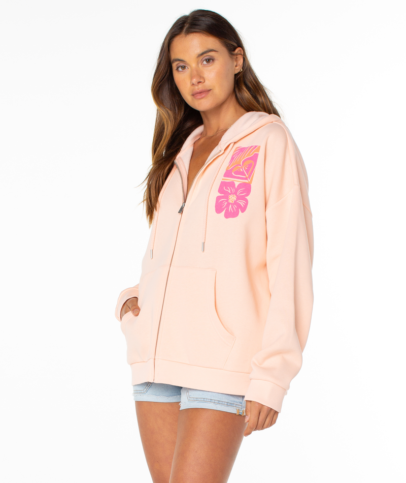 A woman wears the Roxy Evening Lineup Hoodie in light peach with pink floral chest graphics and light denim shorts, standing against a plain white background.