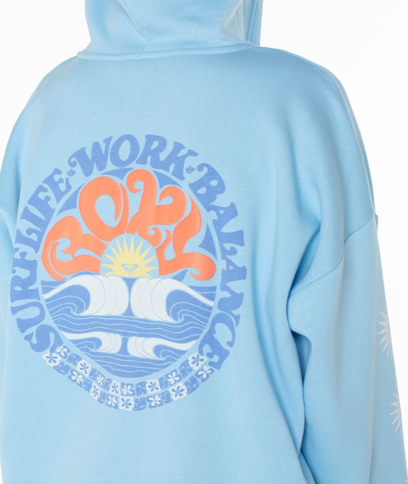 Someone wearing the Roxy Evening Lineup Hoodie in light blue, featuring beach-inspired graphics and a colorful back design with “SURF LIFE WORK BALANCE 2024” above waves, a sunrise, and decorative patterns.