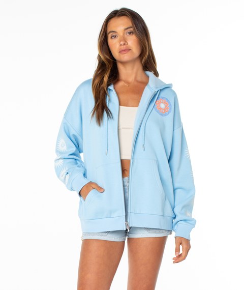 A woman wears the Roxy Evening Lineup Hoodie in light blue, featuring beach-themed graphics, over a white crop top and light denim shorts, standing against a plain white background.